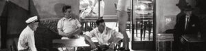 An image in black and white of three casually dressed men sitting outside a closing bar. One of them appears to be a cook, and they are looking around