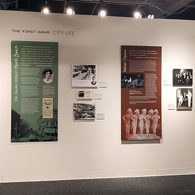 On this wall, there is the title "First Wave: City Life." Below the title, there are two panels: one green panel dedicated to North Beach and another brown panel dedicated to the Opera. Between these two panels, there are three black and white images from the early 1900s.