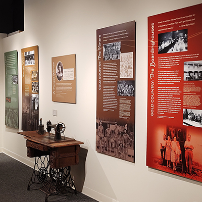 This wall has five panels. The panel on the far left has text that is not readable. The third panel features a sewing machine below it. The remaining two panels are as follows: one in brown covering the topic of mines, and the other covering boarding houses.