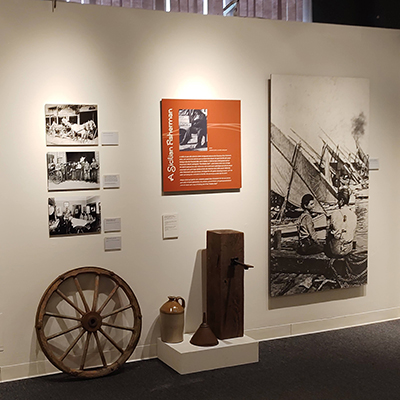 Starting from the left, there are three black and white pictures stacked vertically. In the middle, there is a panel about sailors, and next to that, a nice image of two children at the pier. On the floor, there are several objects: a wheel, a type of water or wine container, and a pillar.