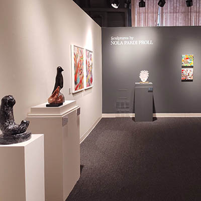 A visual of two walls meeting at a corner. The left wall is white and displays sculptures by Nola Pardi Proll along with artworks by Kara. The dark gray wall further away also features a sculpture by Nola and two smaller paintings by Kara