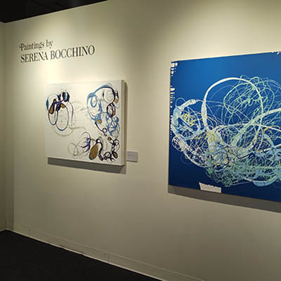 A wall dedicated to Serena Bocchino (her name is in the top right corner). There are two abstract paintings featuring irregular circles of colors. The painting on the left is on a white background, while the one on the right is on a blue background.