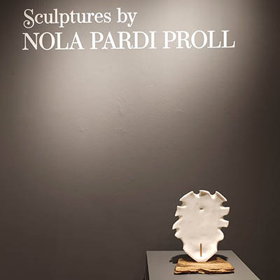 A dark gray wall detail with the name 'Nola Pardi Proll' at the top in white, and one of her white sculptures displayed at the bottom