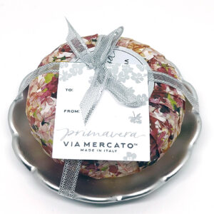 A circular soap wrapped in a warm-hued floral paper, atop a silver dish with a scalloped edged, wrapped in a silver ribbon and "Primavera: Via Mercato" tag.