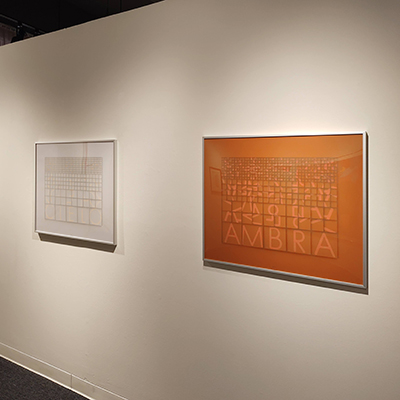 An image of two artworks from the exhibit: one artwork consists of white squares and some text that is not legible, while the other artwork features a series of orange squares with the word "AMBRA" at the bottom of the artwork in a lighter orange