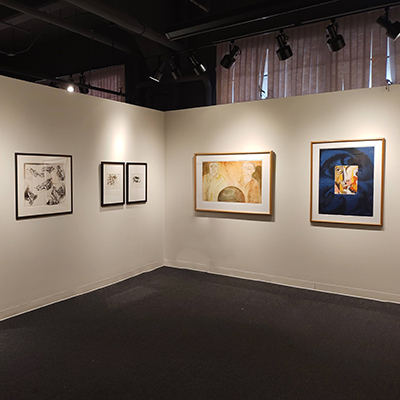 An image showing the corner of two walls. On one wall, there are three black-and-white pencil drawings. On the other wall, there are two artworks: one depicts two men looking at a sphere, and the other is an abstract-style face drawing.