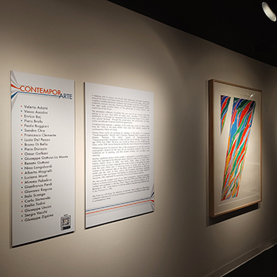 An image of the exhibit wall with two panels: one panel lists the artists, and the other panel provides a description of the permanent collection. To the right side, there is an image of one of the artists with the number 7 depicted in multiple colors