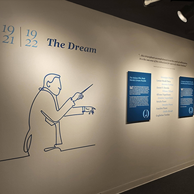 An image of a long white wall. On the left side, there is a silhouette of Merola in dark blue with the date '1921-1922' above it and the title 'The Dream.' On the right side, there are two dark blue panels with the names of the opera founders written in dark blue and light blue in the middle.