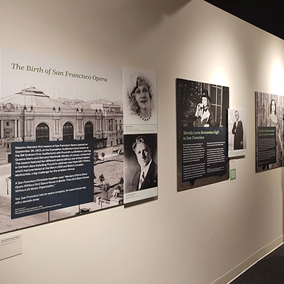 An image of a white wall with several panels showing the history of the birth of the San Francisco Opera. There is an image of an old building where the SF Opera started its official season, followed by black and white images of two opera singers, one man and one woman. These panels are followed by another two panels with images of Beniamino Gigli, and at the end, a black and white image of the singer Claudia Muzio.