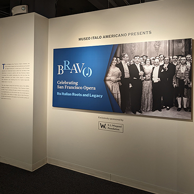 An image to the title wall from the exhibition. It is a long horizontal panel divided in half. On the left side there is the title of the exhibition: Bravo, Celebrating San Francisco Opera. It's Italian Roots and Legacy in White and light blue on a dark blue background. On the right a black and white image from the opera La Traviata in 1932. The picture features a large group of opera singers, including the famous Claudia Muzio and opera director Gaetano Merola. They are all beautifully dressed, and the picture was taken at the end of the show.