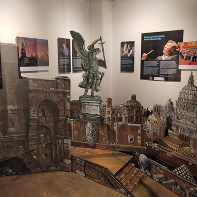 In front of the image, there is a detail of the Tosca scenography model. In the background, slightly out of focus, are several panels showing opera singers and opera directors over the years.