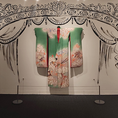 An image of a white wall with a black drawing of the SF Opera stage. In the middle, there is a real dress hanging from the wall. The dress is a kimono used in a performance of Madama Butterfly. The kimono is green with cherry blossom flowers in pink and white, and it features some details of Japanese culture.