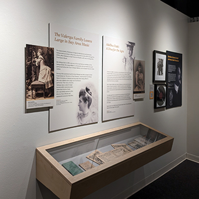 An image of a white wall with several panels. The panels closest to the viewer are about Ida Valerga, an opera singer. There is an image of Valerga kneeling on a chair and another one of her profile. We also see another female opera singer, depicted in both a portrait and a 3/4 body image. Following these is a black and white image, a record, and another panel on a dark gray background with a profile image.