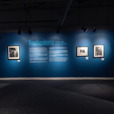 One of the exhibit walls has a dark blue background with the word "Neorealismo" in a lighter blue. Below, there are two columns of text. On each side of the text, there are black-and-white images: one on the left side and two on the right side.