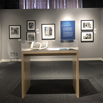 A grey wall with six black-and-white images and a blue text panel, all in the background. In front of the wall, in the middle of the room, there is a display case (vitrine) with books inside.