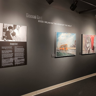 The dark gray wall features a panel with Giovanni Cerri's bio and the title "Giovanni Cerri: 2020: Milan in the Hour of the Wolf." To the right of the panel, there are two paintings: one depicting an orange tram and the other showing a lady with a COVID mask.