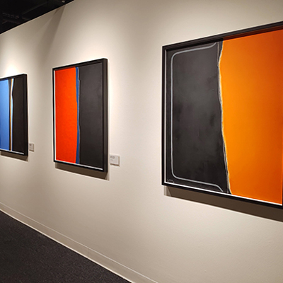 On this white wall, there are three abstract paintings. Starting from the left: The first painting is half blue and half black, divided by a thin blue line. The second painting is red and black, divided in the middle by a blue line. The last painting on the right is black and orange, with a white line in the middle.