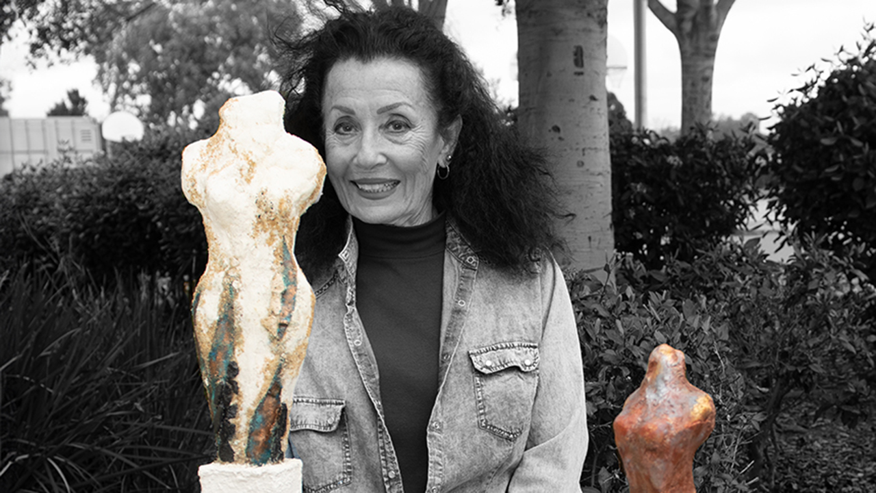 An image in black and white of Tricia Grame standing between two stylized sculptures of women in color. Tricia has long hair and is casually dressed