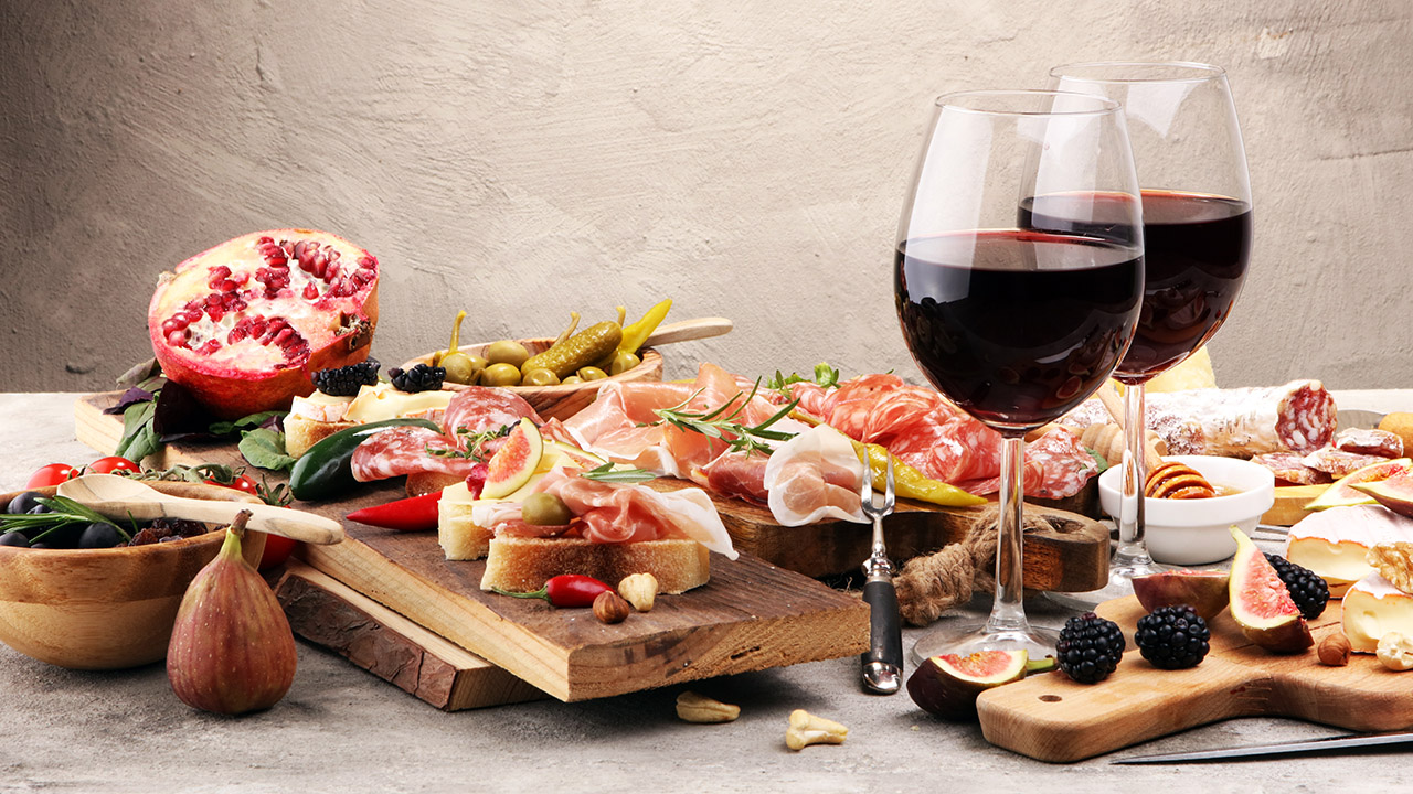 An image of a plate full of appetizer foods: prosciutto, bread, figs, cheese, salami, blackberries, and pickles. In the middle, there are two glasses full of red wine.