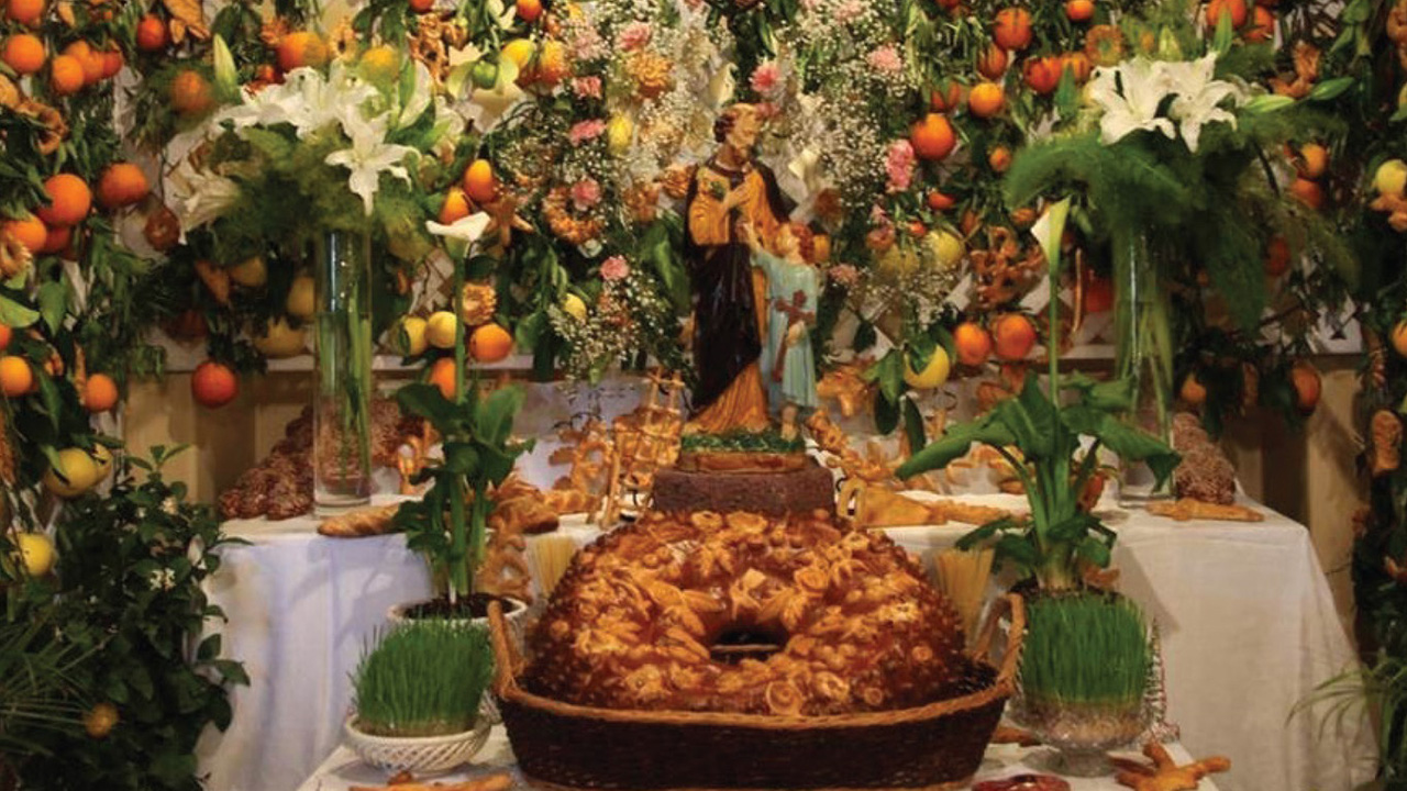 On a background of lemon and orange plants, there is an altar with food on both sides and two vases with white flowers on each side. In the middle of the table is a sacred sculpture of a saint with a child, and in the front, a cake.