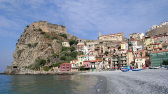 An image of the beach in Scilla, a town in Calabria, Italy. The beach is in the foreground, with many houses crowded on a rocky cliff in the distance, and the Ruffo Castle overlooking the scene.