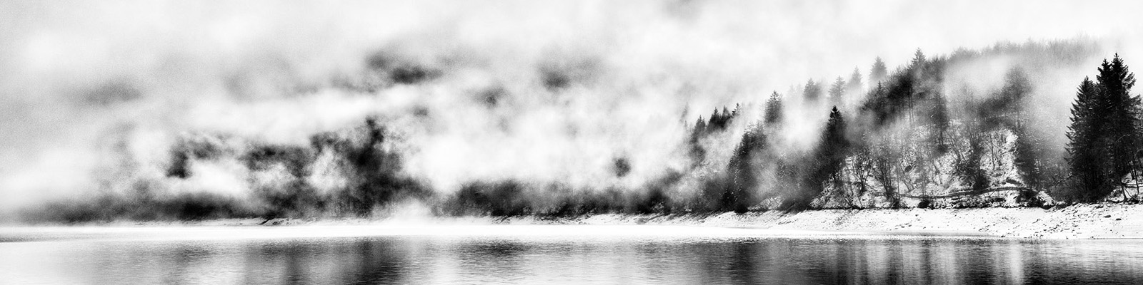 A black-and-white image of a lake in winter with snow on the banks and low clouds covering much of the sky.