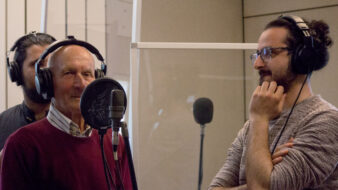 A recording session featuring an elderly man singing into a microphone, wearing headphones, while two younger men listen attentively in a professional studio setting.