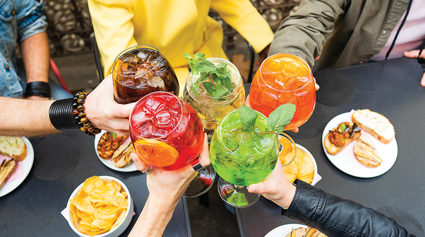 Two arms protrude from the foreground, one wearing rings and holding a red cocktail, the other wearing a leather jacket and holding a green cocktail. Three people sit in the background, their torsos (in blue, yellow, and green clothing) and arms visible. One holds a brown cocktail, the other a clear cocktail, and the third an orange cocktail. All five glasses of cocktails are clinked together over two black tables holding small bites like potato chips and bread with grilled vegetables.