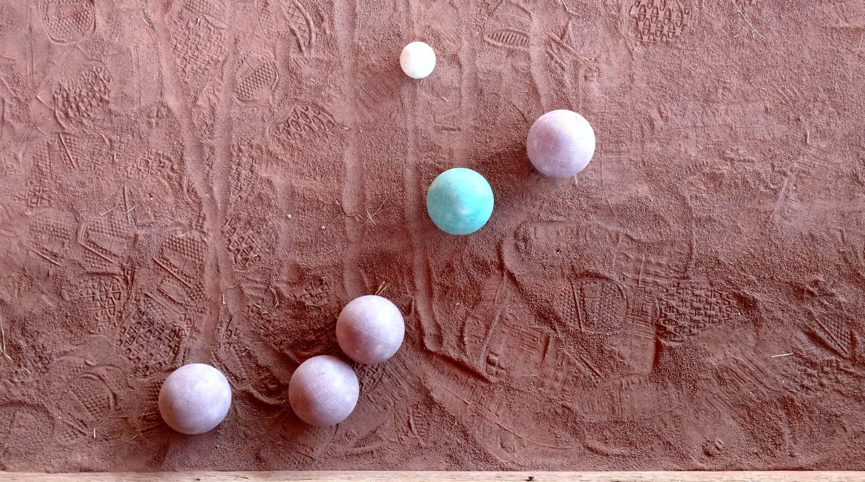 Six balls against a packed sand background. At the lower left of the photo are three pink balls, in the middle center is a blue ball, and just to the right of the blue ball is another pink ball. There are all the same size. Above the blue ball is a smaller white ball. There are multiple footprints in the sandy dirt.