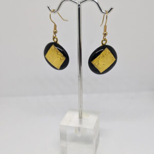 A pair of earrings with circular glasswork featuring gold foil square against a black background, on gold hooks.