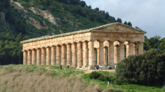 Photograph of the ruins of a temple in Sicily surrounded by greenery and a mountain in the background. The columns and pediment are still intact. People are also shown walking around the temple.