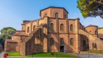 A photograph of St. Vitale, a church in Emilia Romagna. The church is a brick structure with a hexagonal base with two levels and six windows on each side. Above there is a smaller hexagonal third level. The church has flying buttresses coming of the sides and differently sized turrets.