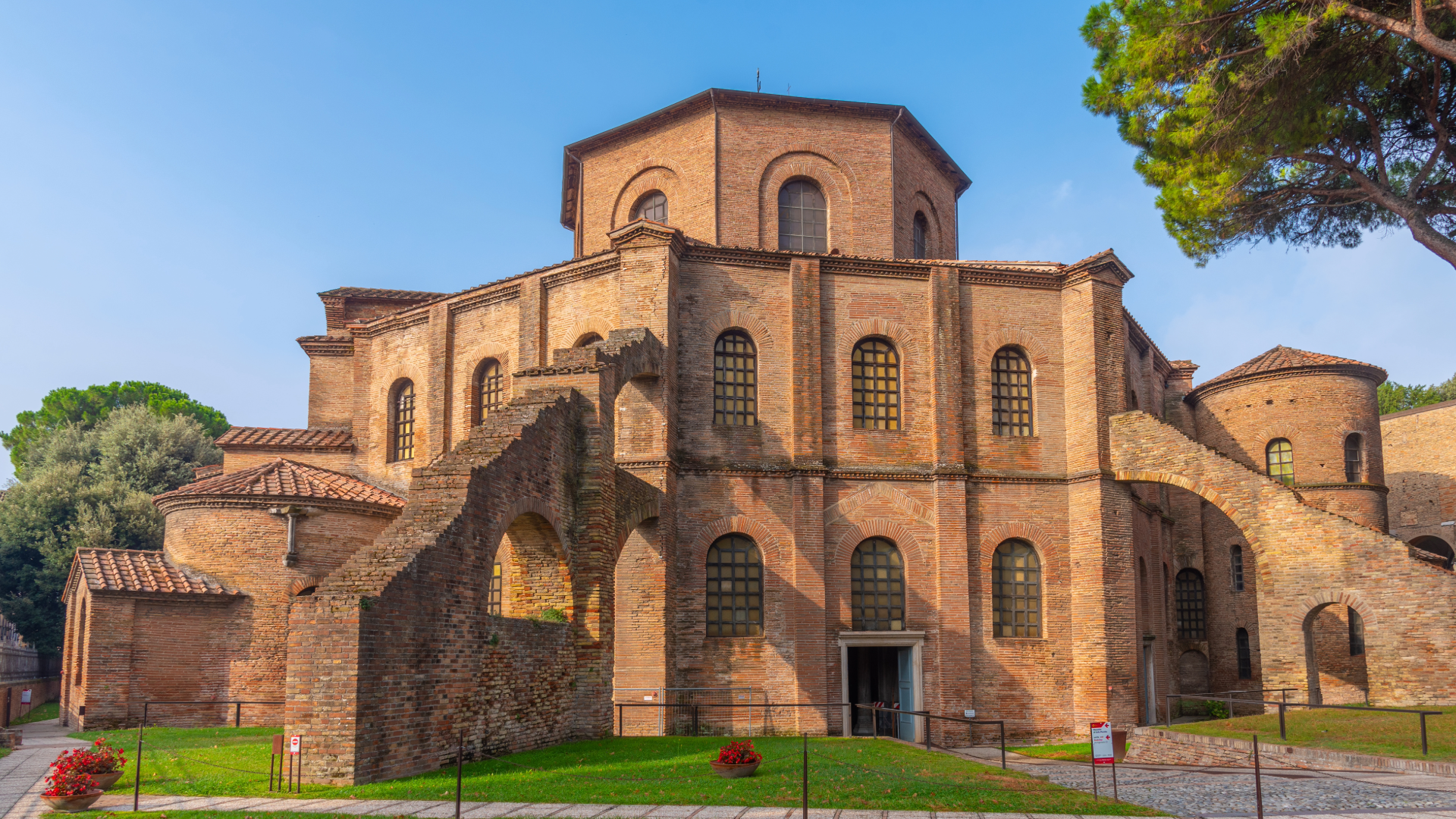 A photograph of St. Vitale, a church in Emilia Romagna. The church is a brick structure with a hexagonal base with two levels and six windows on each side. Above there is a smaller hexagonal third level. The church has flying buttresses coming of the sides and differently sized turrets.
