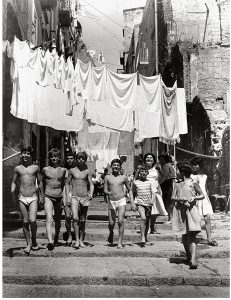 A black-and-white image of several young men in underwear. At the top of the image, many white clothes are hanging from wires strung between two houses.