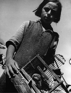 A black-and-white image of a child in poor clothing. The child is carrying a cage with an animal inside.