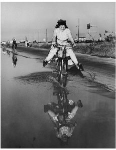 An image in black and white of a girl riding a bicycle into a puddle