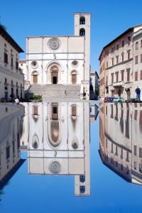 The image shows a church facade with its reflection in the water in the lower half of the image. The facade of the church is white, featuring a large door in the middle and two smaller doors on the sides.