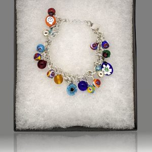 A charm bracelet is being crafted using the traditional millefiori (thousand-flowers) technique, where thin rods of glass (called murrine) are carefully arranged in a pattern and then heated until they fuse together. The bracelet is designed in bright colors reminiscent of carnevale.