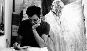 A black and white image shows Giovanni Cerri drawing in his studio, with a painting of Pope Francis in the background.